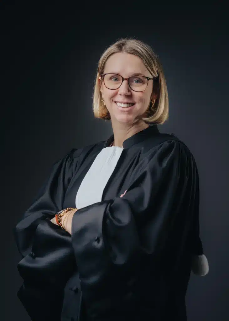 Portrait d'une avocate en robe noire avec rabat blanc Avocate souriante à lunettes en robe noire avec rabat blanc, bras croisés sur fond sombre.